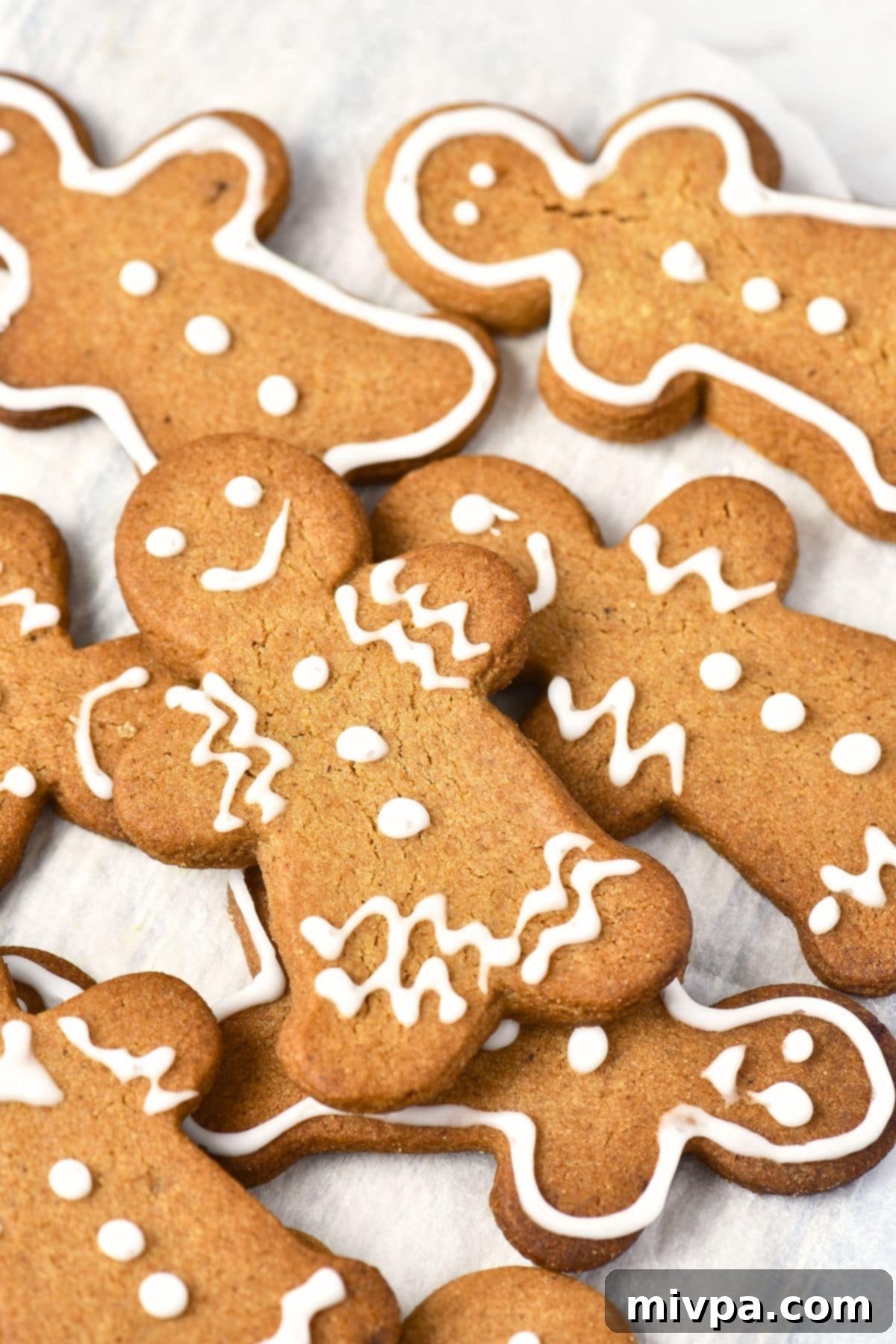 Gingerbread Men Recipe (Gluten-Free, Dairy-Free) Up close view of gingerbread men cookies stacked on parchment-lined sheet, showcasing intricate icing details.