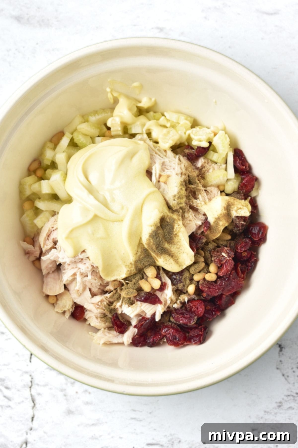 Chicken Salad with Cranberries A bowl with ingredients for chicken salad and dried cranberries, with dressing being poured over.