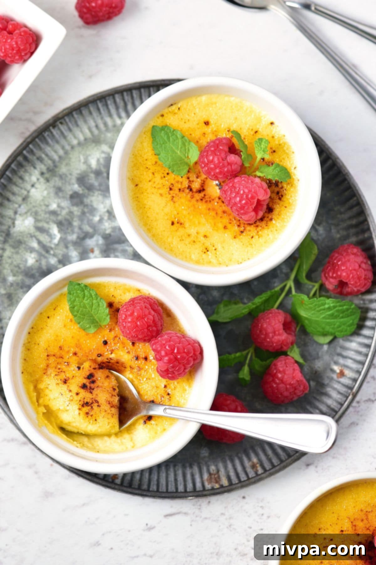 A close-up of a crème brûlée in a ramekin, garnished with fresh berries and mint, ready to be served on a white background.