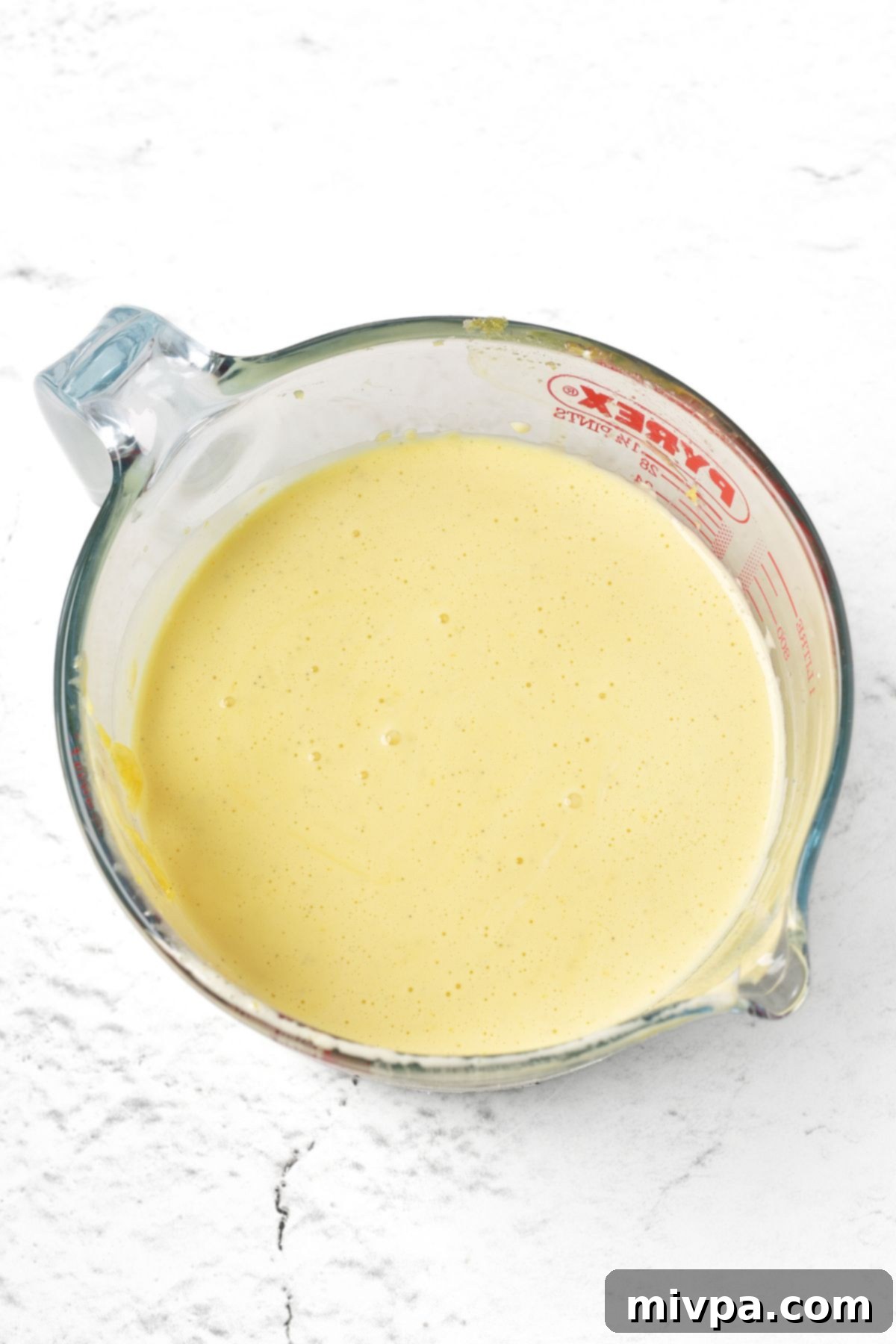 Warm cream being slowly poured into a bowl of whisked egg yolks and sugar, while stirring constantly to temper the eggs.