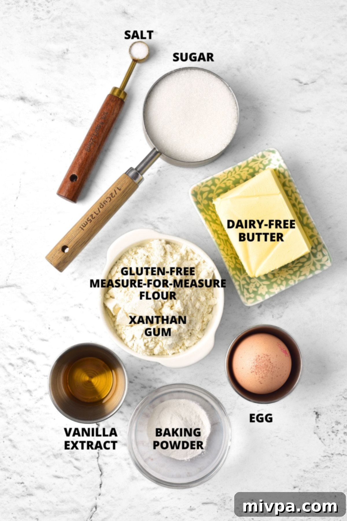 A carefully arranged display of all the ingredients needed for gluten-free Christmas sugar cookies, including dairy-free butter, sugar, gluten-free flour, egg, vanilla extract, baking powder, xanthan gum, and salt, laid out on a clean marble board.