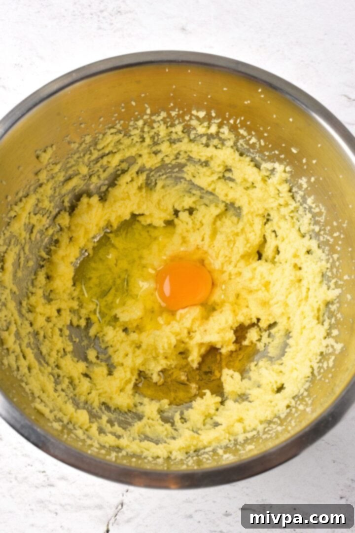 A raw egg cracked into a bowl of yellow, creamed butter and sugar dough, illustrating the 'Add Egg' step in the cookie-making process.