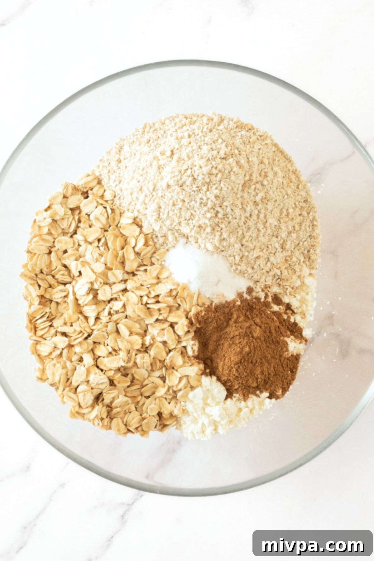 How to Make Gluten Free Oatmeal Muffins A glass bowl containing a mix of dry ingredients: rolled oats, gluten-free flour, ground cinnamon, and baking powder.