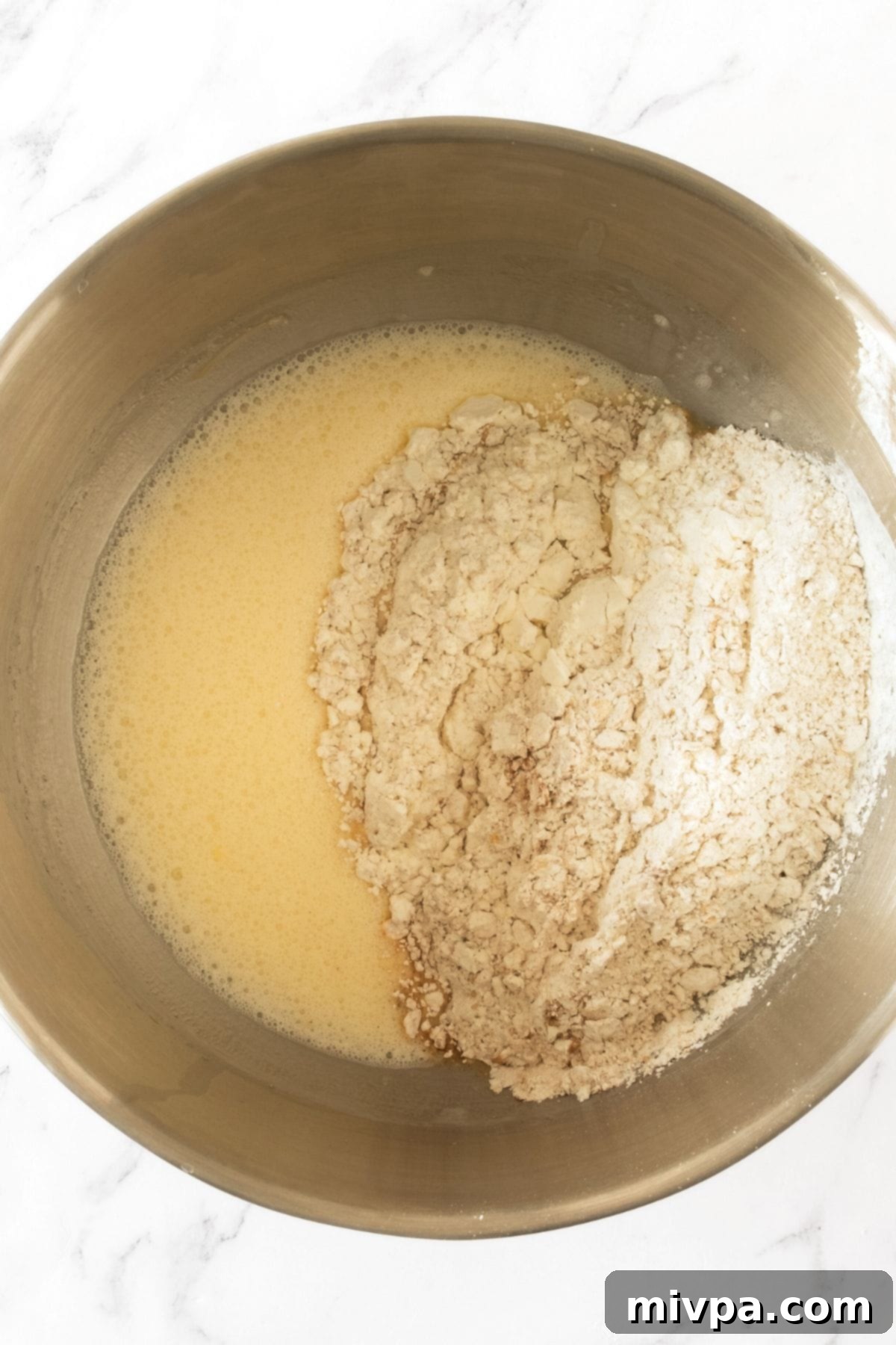 Gluten Dairy-Free Oatmeal Muffins Dry flour mixture being added to the pale yellow liquid mixture in a metal bowl, illustrating the combining step.