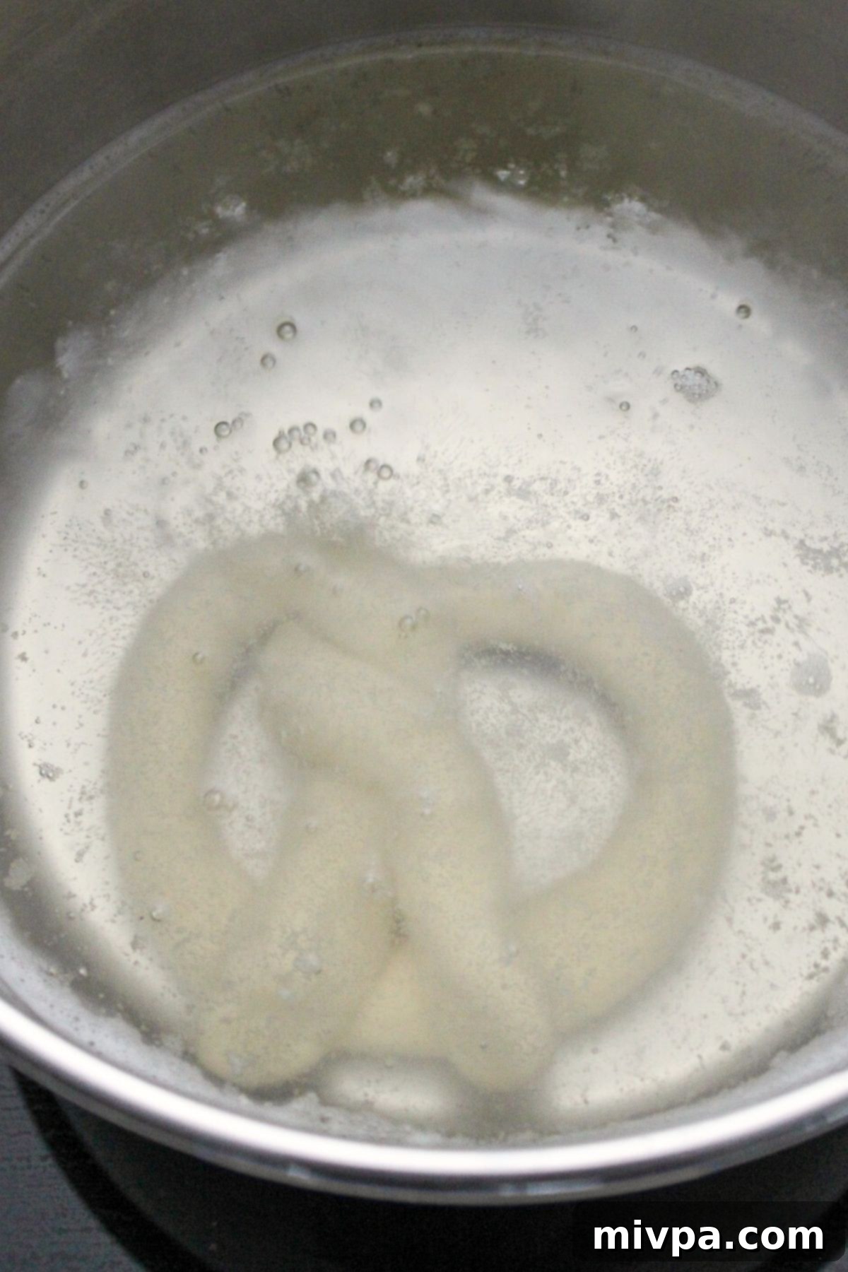 A shaped gluten-free pretzel being carefully boiled in a simmering soda bath in a pot on the stove, crucial for its chewy texture.