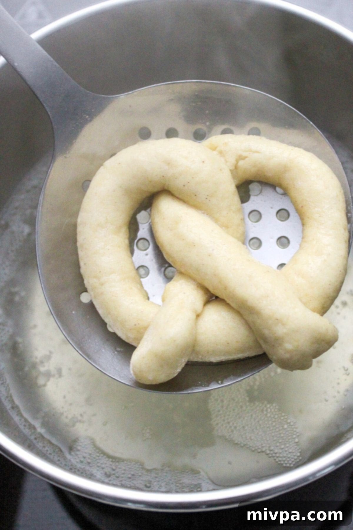A slotted spoon carefully lifting a boiled gluten-free pretzel from the soda bath, demonstrating the removal process before baking.