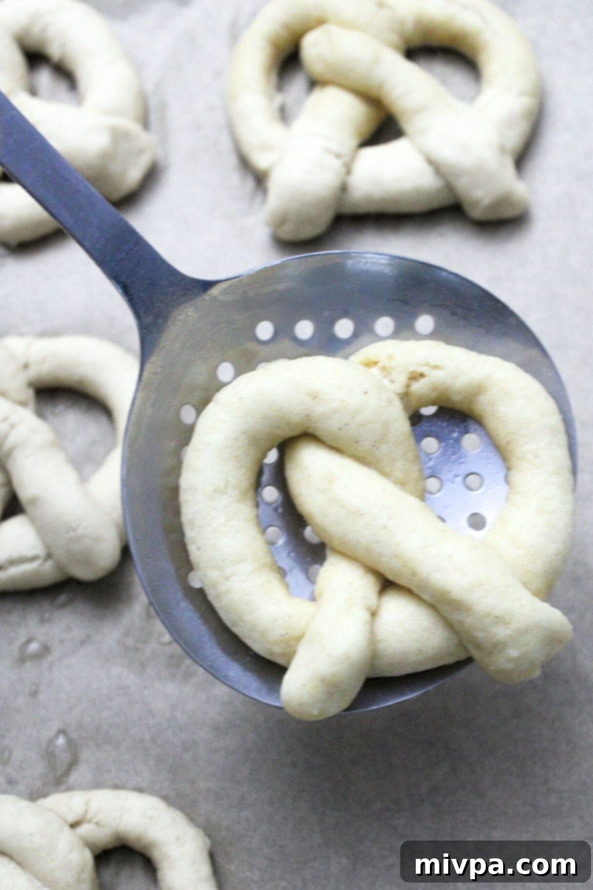 Boiled gluten-free pretzel dough resting on a slotted spoon, ready to be transferred to a baking sheet.