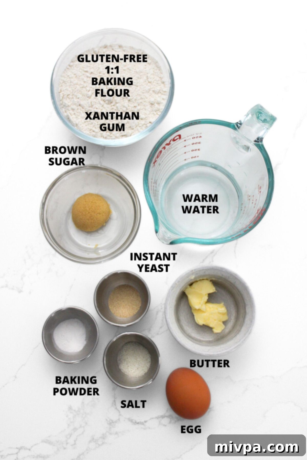 Various ingredients for gluten-free soft pretzels recipe, including flour, sugar, yeast, and butter, neatly laid out on a white board.