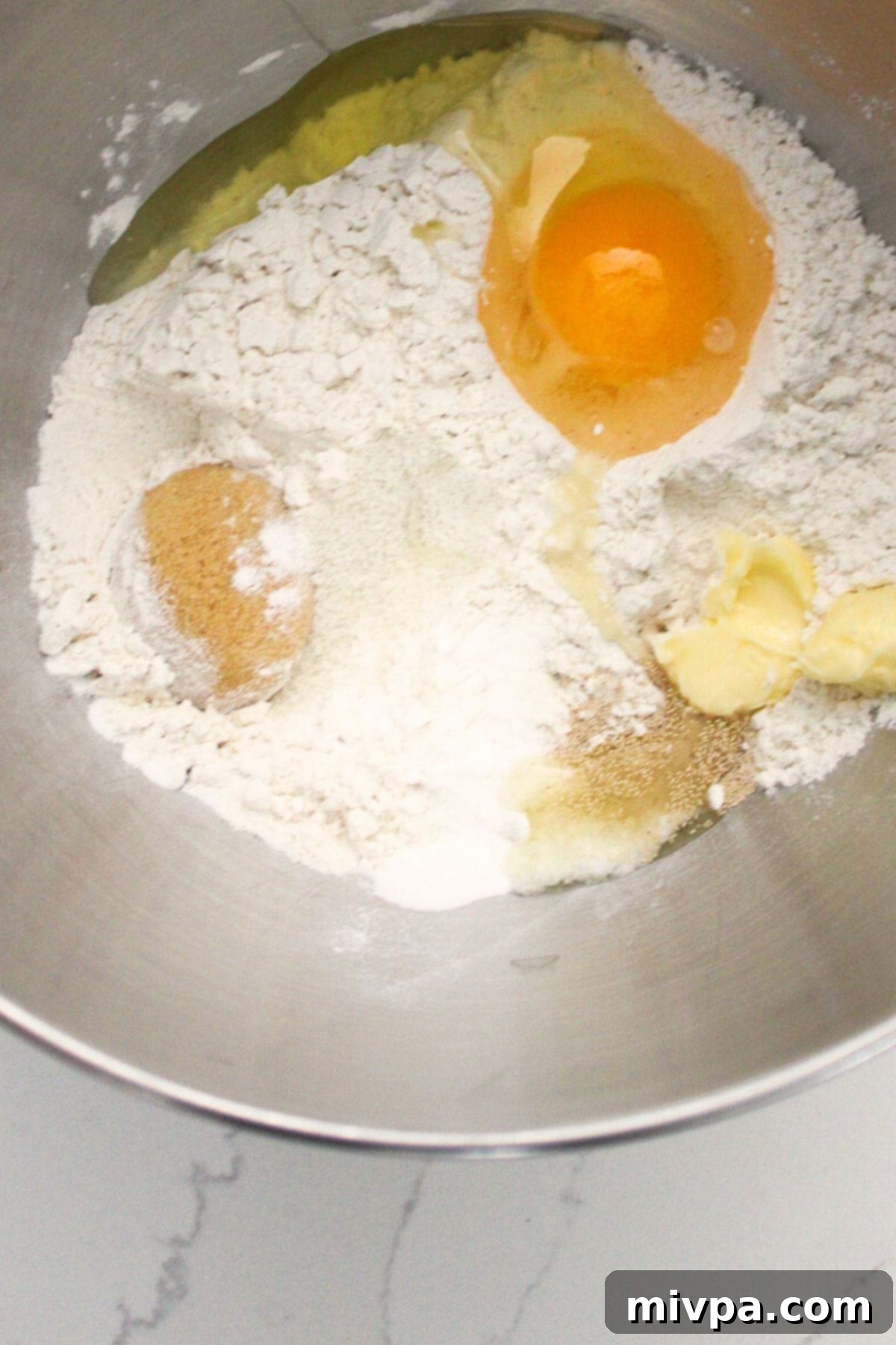 Dry ingredients like gluten-free flour, sugar, and yeast mixed with wet ingredients like egg and butter in a metal mixing bowl, ready to form the pretzel dough.