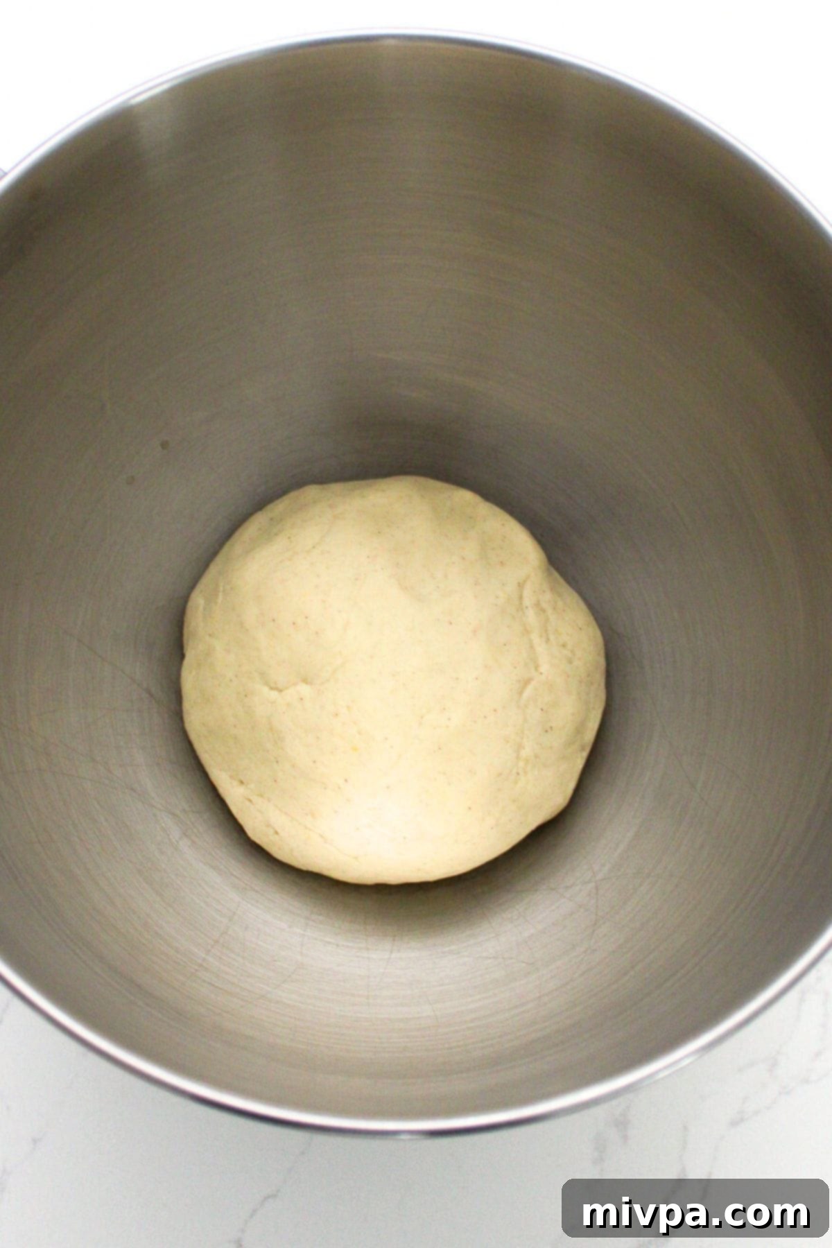A perfectly shaped ball of pretzel dough resting in a metal bowl, covered with plastic wrap, ready for its rising period.