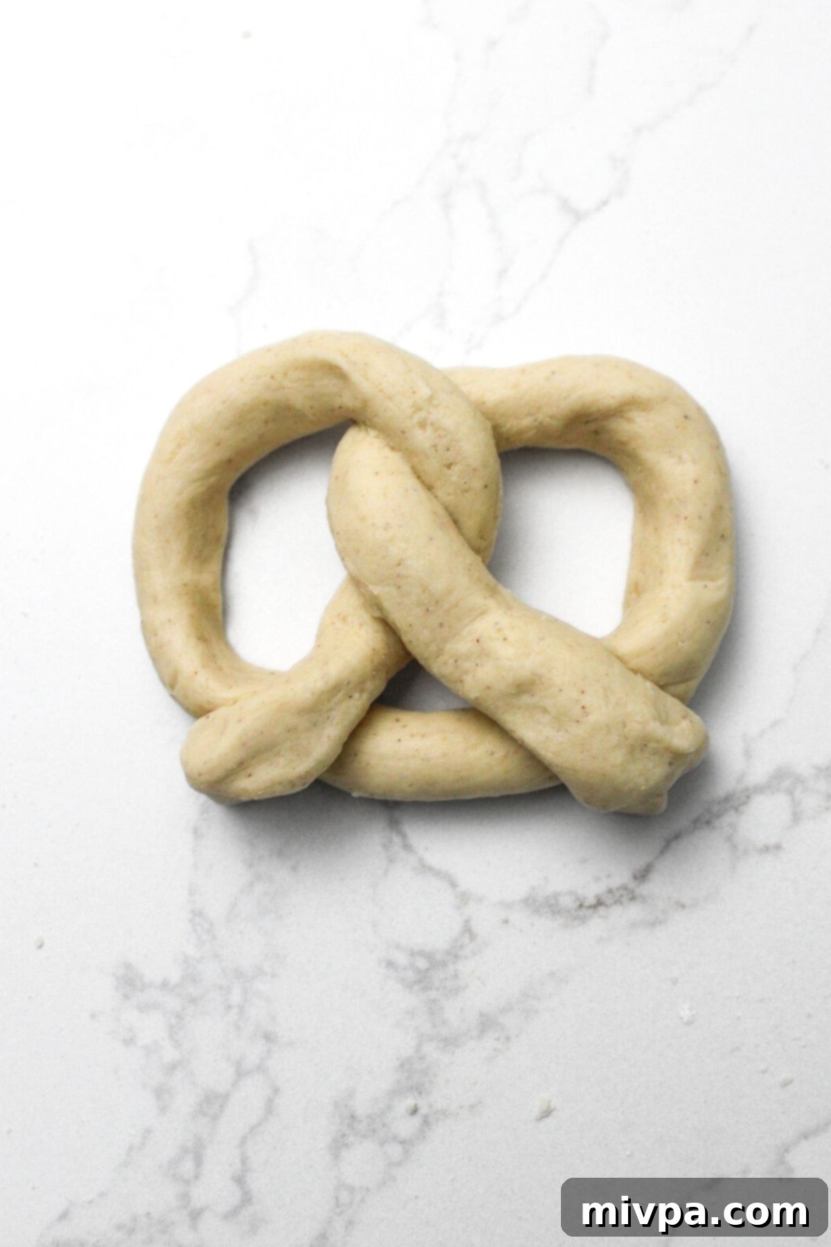 Gluten-free dough fully shaped into the classic pretzel knot on a marble board, ready for the next step in the baking process.