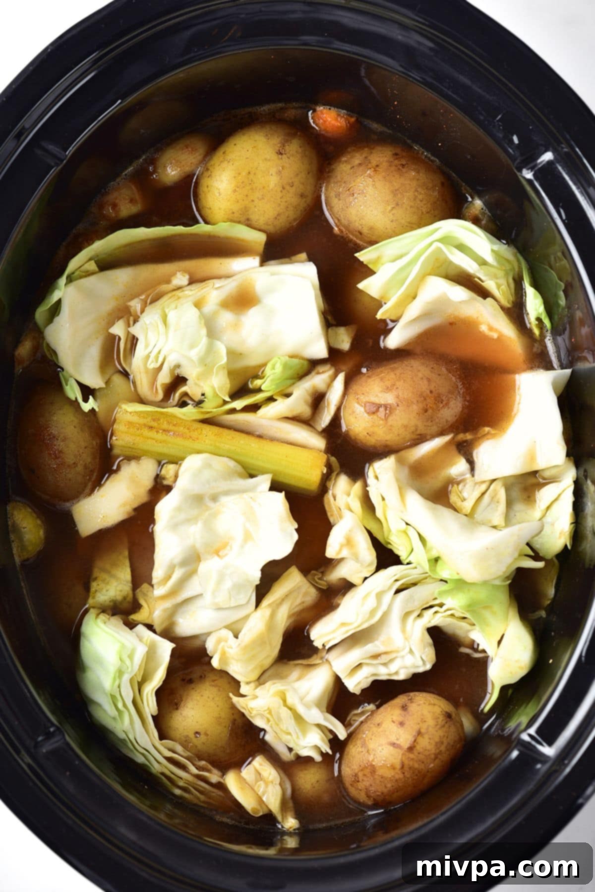 Chopped cabbage being added to the slow cooker with the simmering corned beef and vegetables.