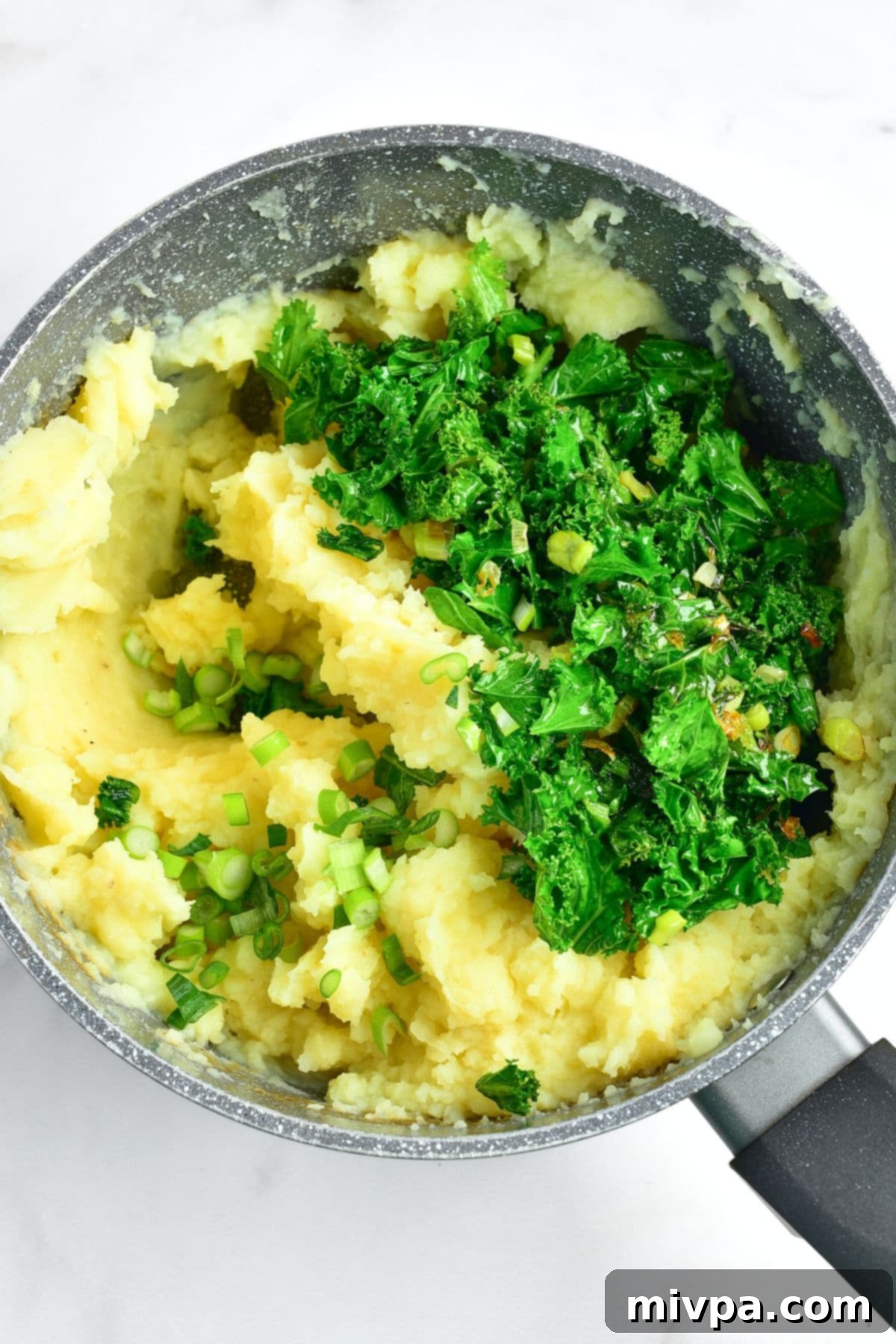Adding Sautéed Veggies to Mashed Potatoes Sautéed kale and green onions being added on top of a pot of mashed potatoes