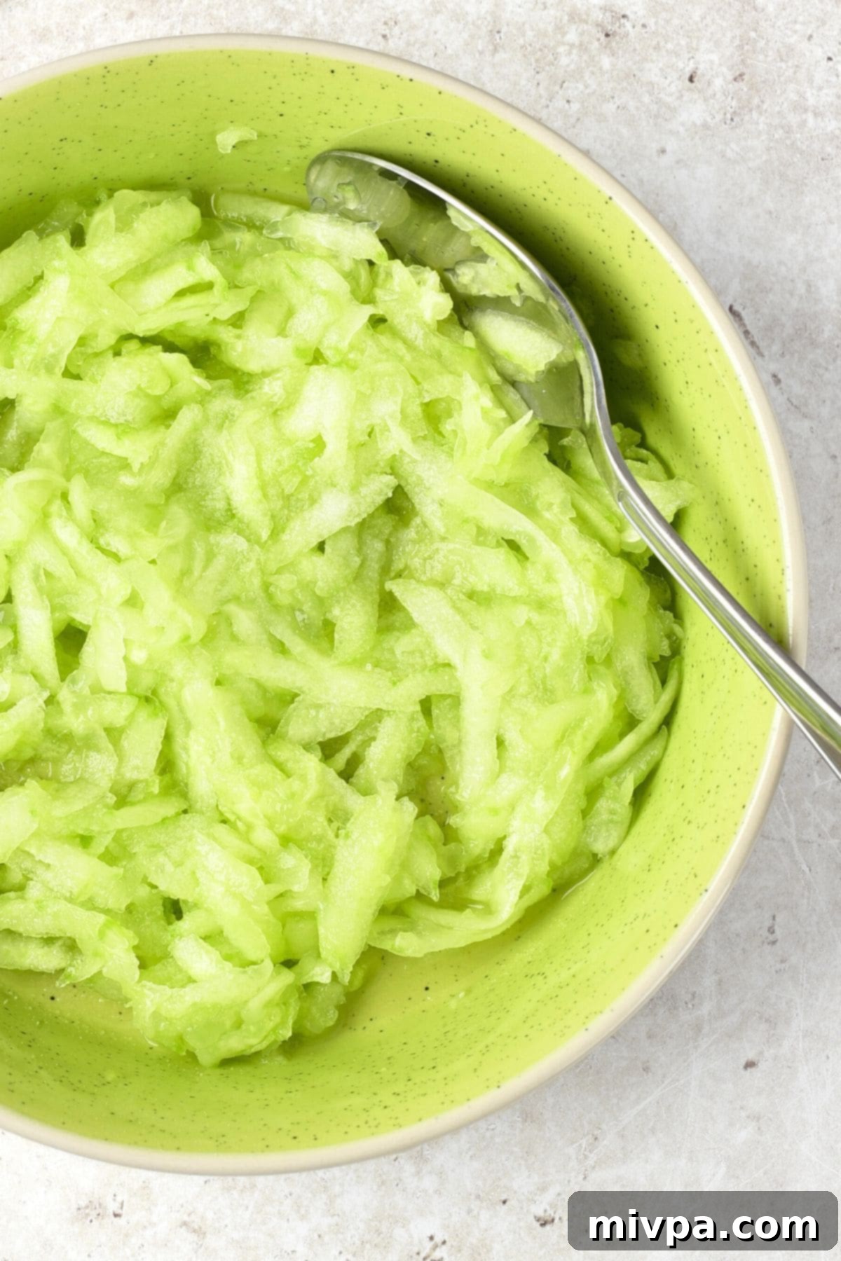 A spoon in a bowl of freshly grated cucumber, sprinkled with salt.