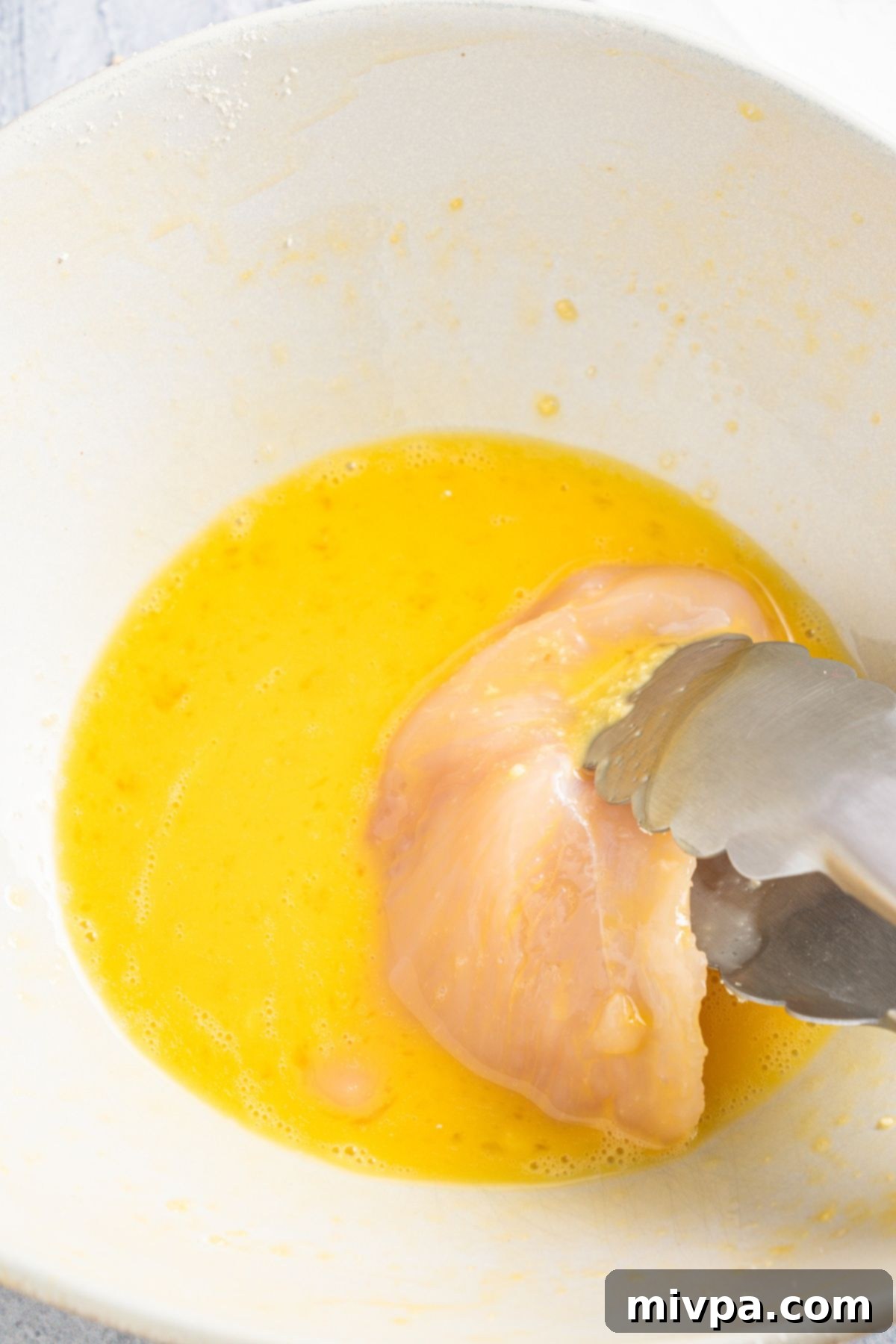 Using kitchen tongs to carefully dip a chicken fillet into a bowl of beaten eggs, ensuring it's thoroughly coated for the breading process.