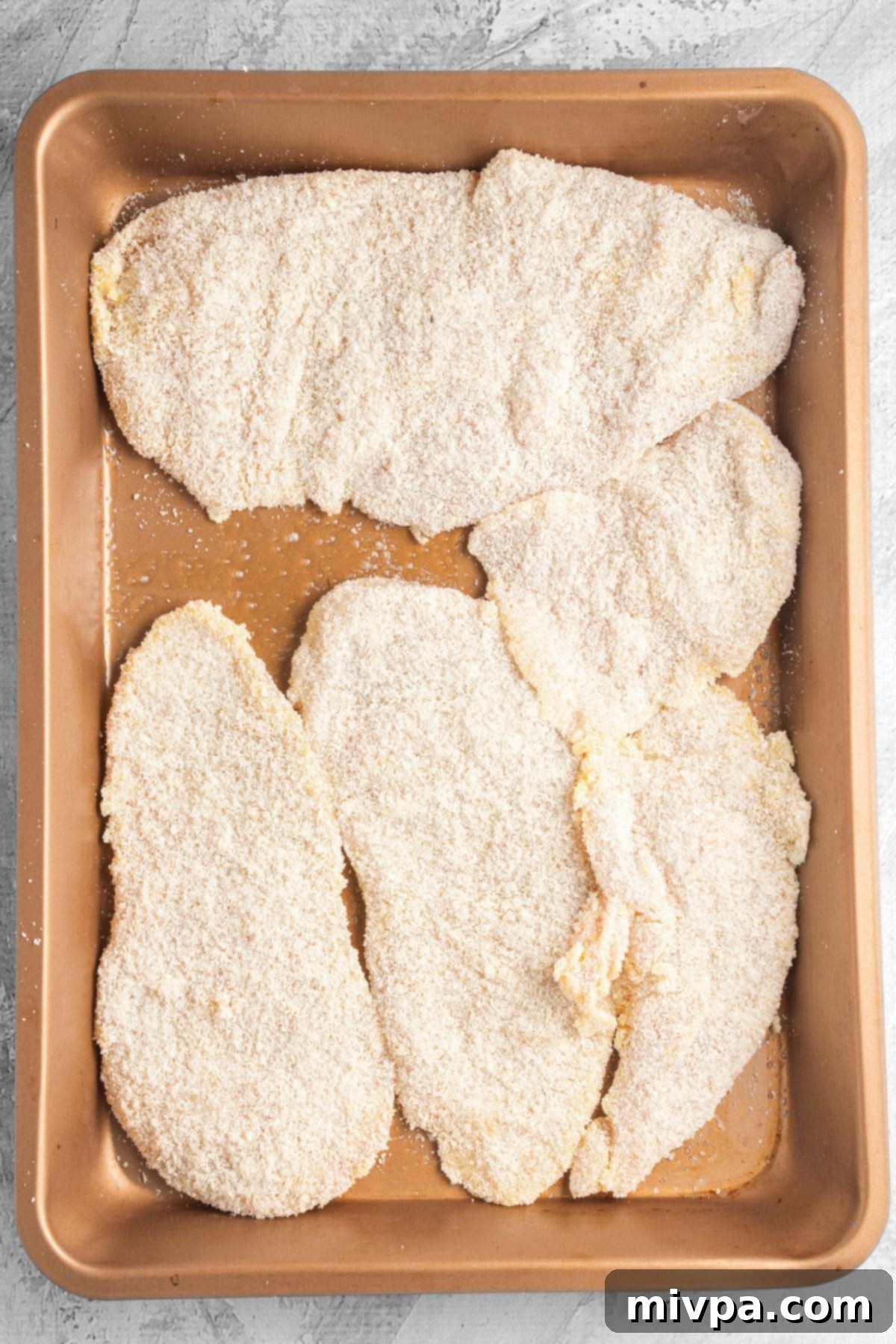 Gluten-free breaded chicken cutlets arranged on a sheet pan, drizzled with oil, and ready to be placed into the oven for baking.