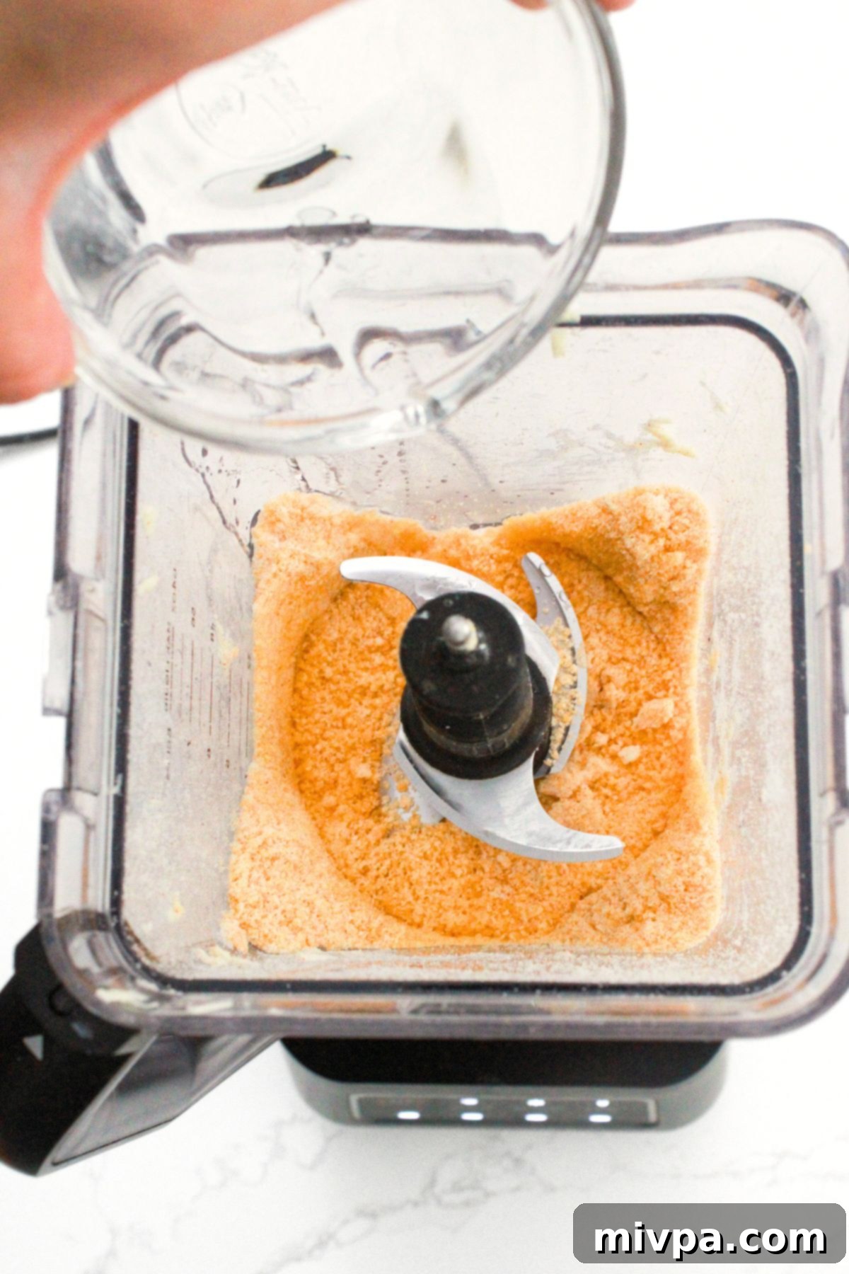 How to Make Goldfish Crackers Ice-cold water being carefully poured into the crumbly orange flour and cheese mixture inside an electric blender, initiating dough formation.