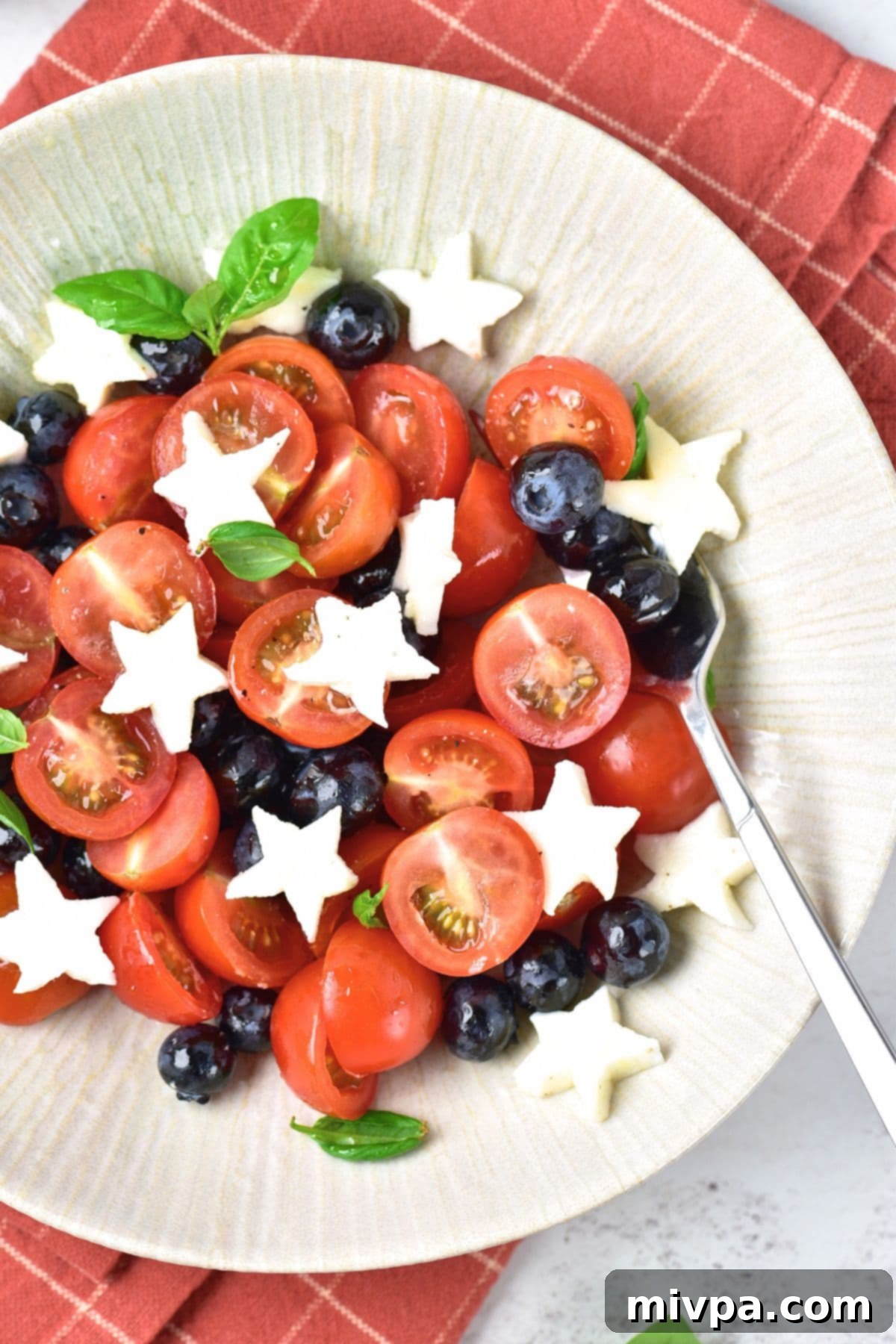 Red, White and Blue Caprese Salad (Gluten-Free) Top down view of sliced tomatoes, blueberries, basil leaves and star shaped mozzarella in bowl.