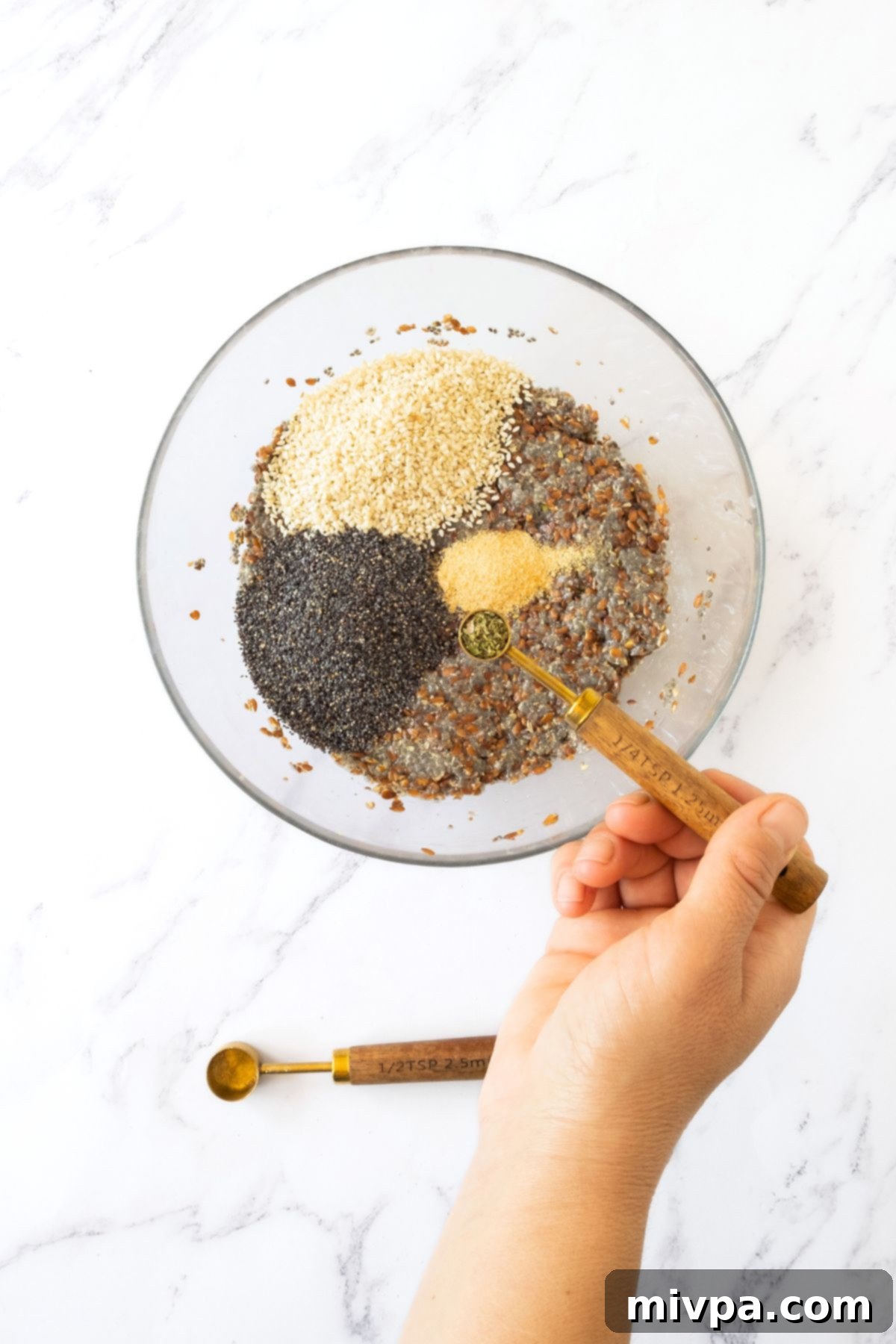 Effortless Gluten-Free Vegan Flaxseed Crackers 2 Adding fragrant dried oregano to a bowl already containing hydrated chia and flax seeds, poppy seeds, garlic powder, and sesame seeds.