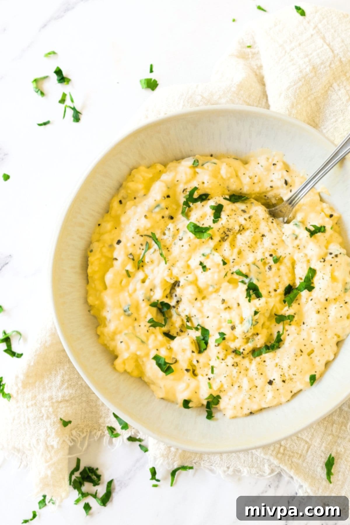 A spoon in a bowl of creamy egg salad topped with chopped parsley.