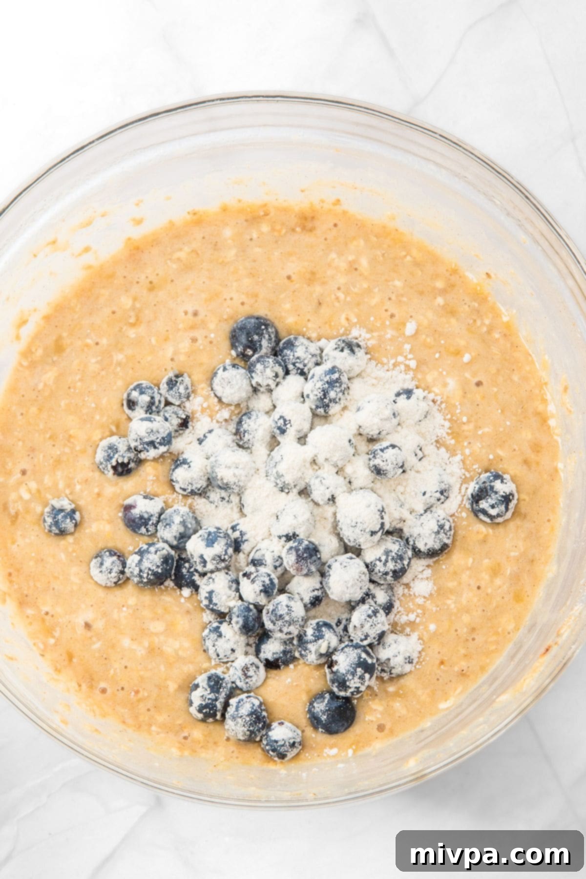 Flour-coated blueberries on top of muffin batter in a bowl, prepared for folding.