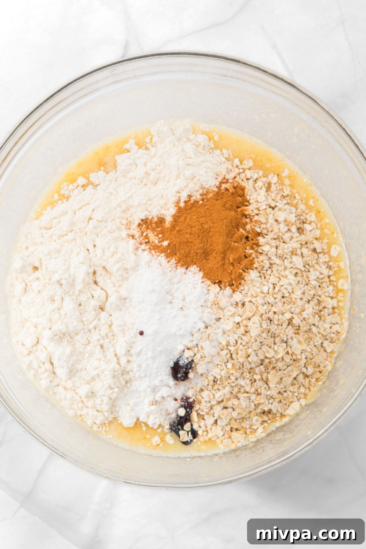 A bowl with oats, flour, cinnamon, baking powder and pale yellow liquid, illustrating the dry ingredients being added.