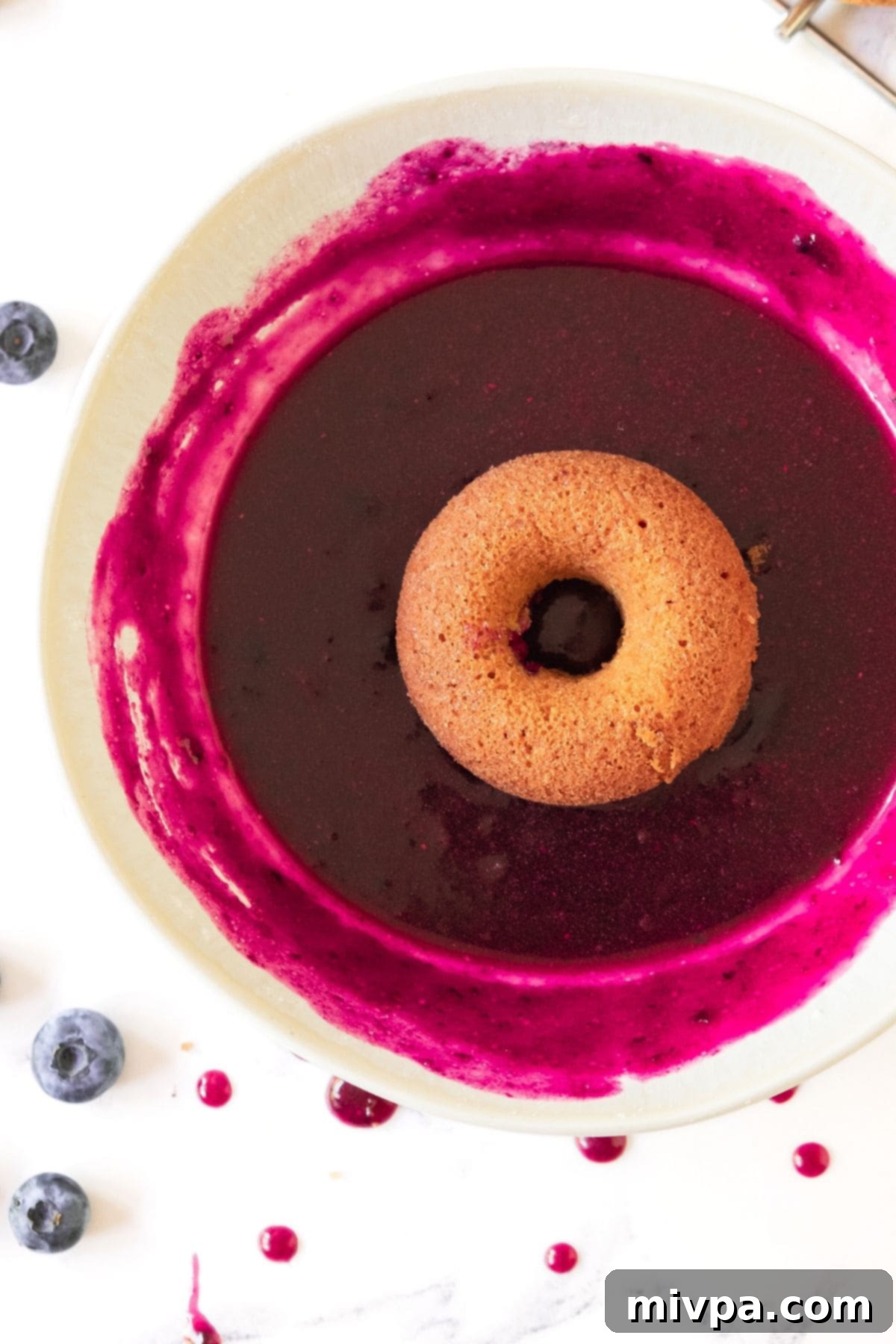 Homemade Blueberry Glaze (Gluten-Free, Vegan) A donut in a bowl of homemade blueberry glaze, showcasing its vibrant purple hue and glossy texture.