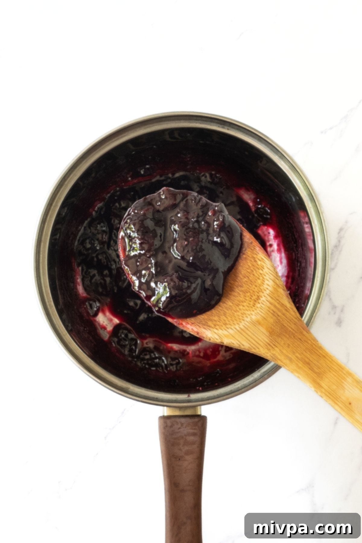 Gluten-Free Vegan Blueberry Glaze Step 1 A spoonful of cooked blueberry mixture being held up, showing its slight thickening and rich color, fresh from the pot.
