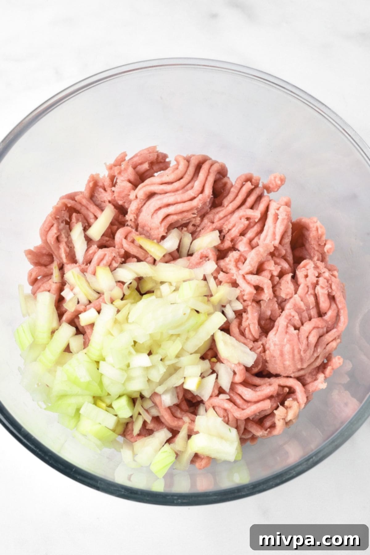 A glass mixing bowl containing finely diced onions combined with raw ground turkey, ready for seasoning.