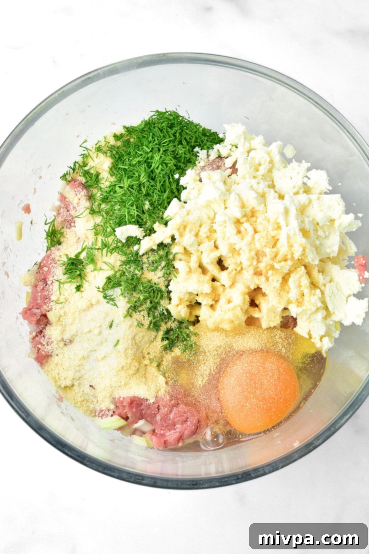 A bowl showcasing ground turkey, almond flour, freshly chopped dill, crumbled feta cheese, and an egg, all ready to be mixed for meatballs.
