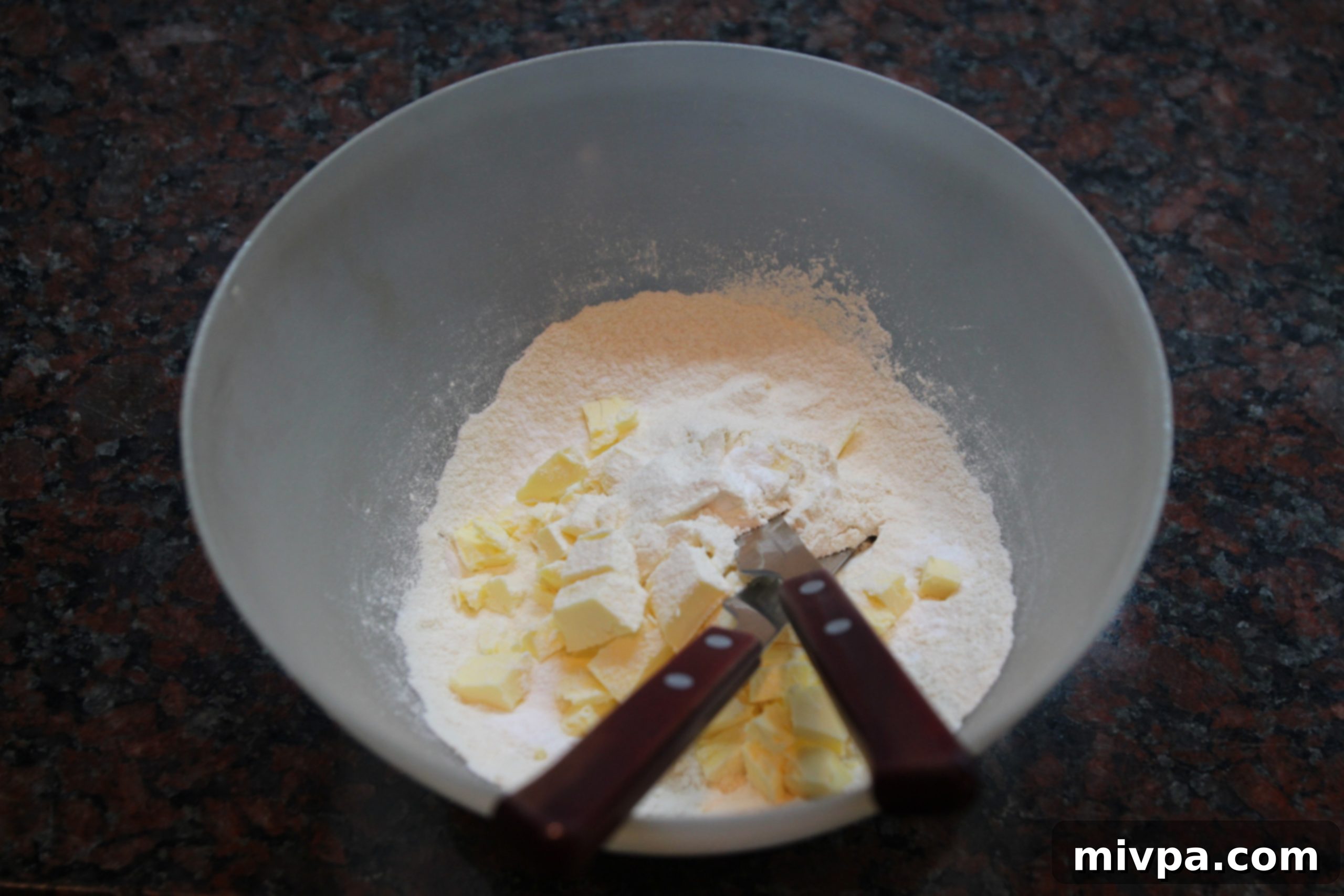 Process of mixing flour, cold butter, and icing sugar in a bowl using two knives, creating a crumbly texture.
