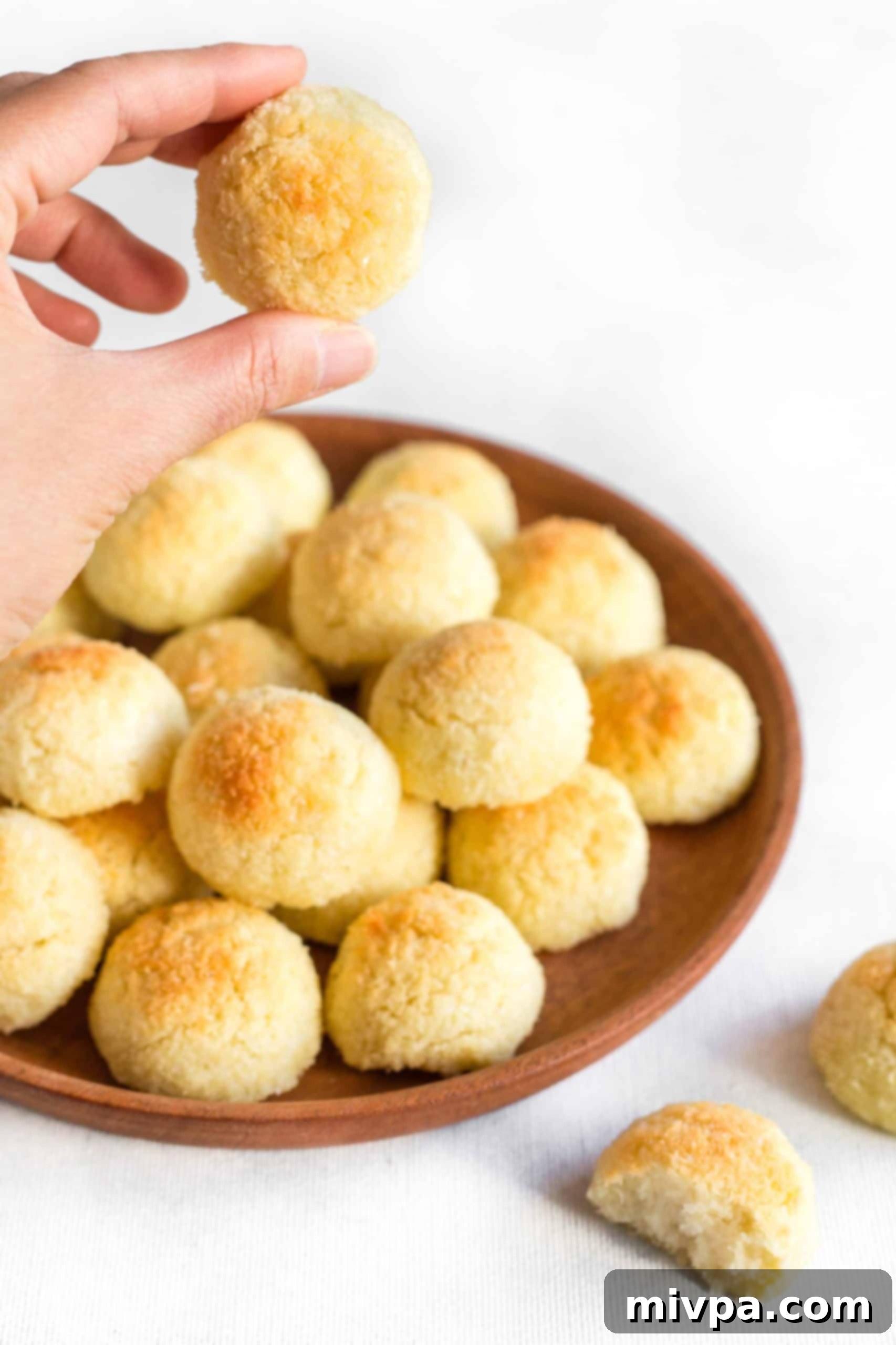 Gluten-Free Dairy-Free Coconut Macaroons - Hand Holding a Cookie A hand gently holding a single gluten-free coconut cookie, with a wooden plate full of more cookies in the background.