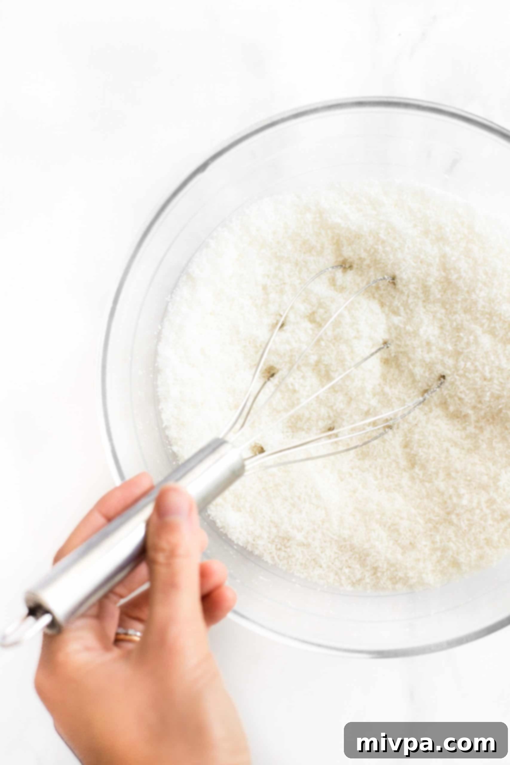 Recipe for Coconut Macaroons (Gluten-Free, Dairy-Free) - Mixing Dry Ingredients A hand is shown whisking dry ingredients, specifically shredded coconut and sugar, in a clear glass mixing bowl.