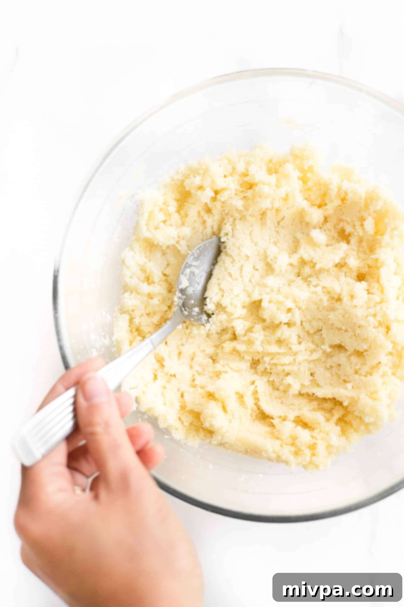 Gluten and Dairy-Free Coconut Macaroons - Dough Mixing A hand uses a metal spoon to mix the wet and dry ingredients, forming coconut cookie dough in a clear glass bowl.
