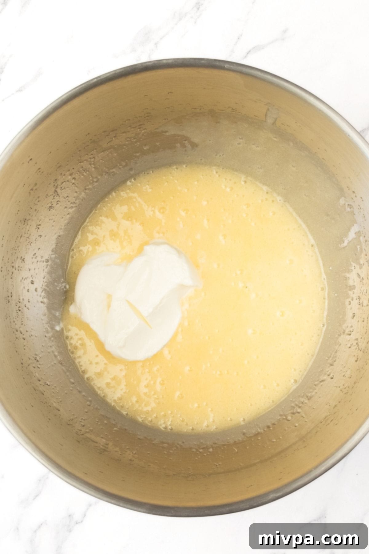 Mixing Wet Ingredients for Easy Swedish Apple Cake Sour cream added on top of a pale yellow cake batter in a mixing bowl, prior to mixing.