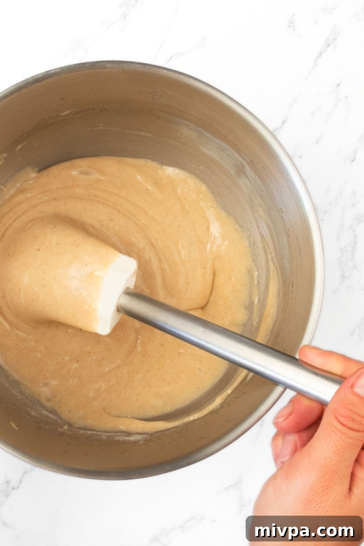 Mixing Batter for Swedish Äppelkaka (Gluten-Free) Mixing cake batter in a large metal bowl with a spatula, creating a smooth, homogeneous texture.