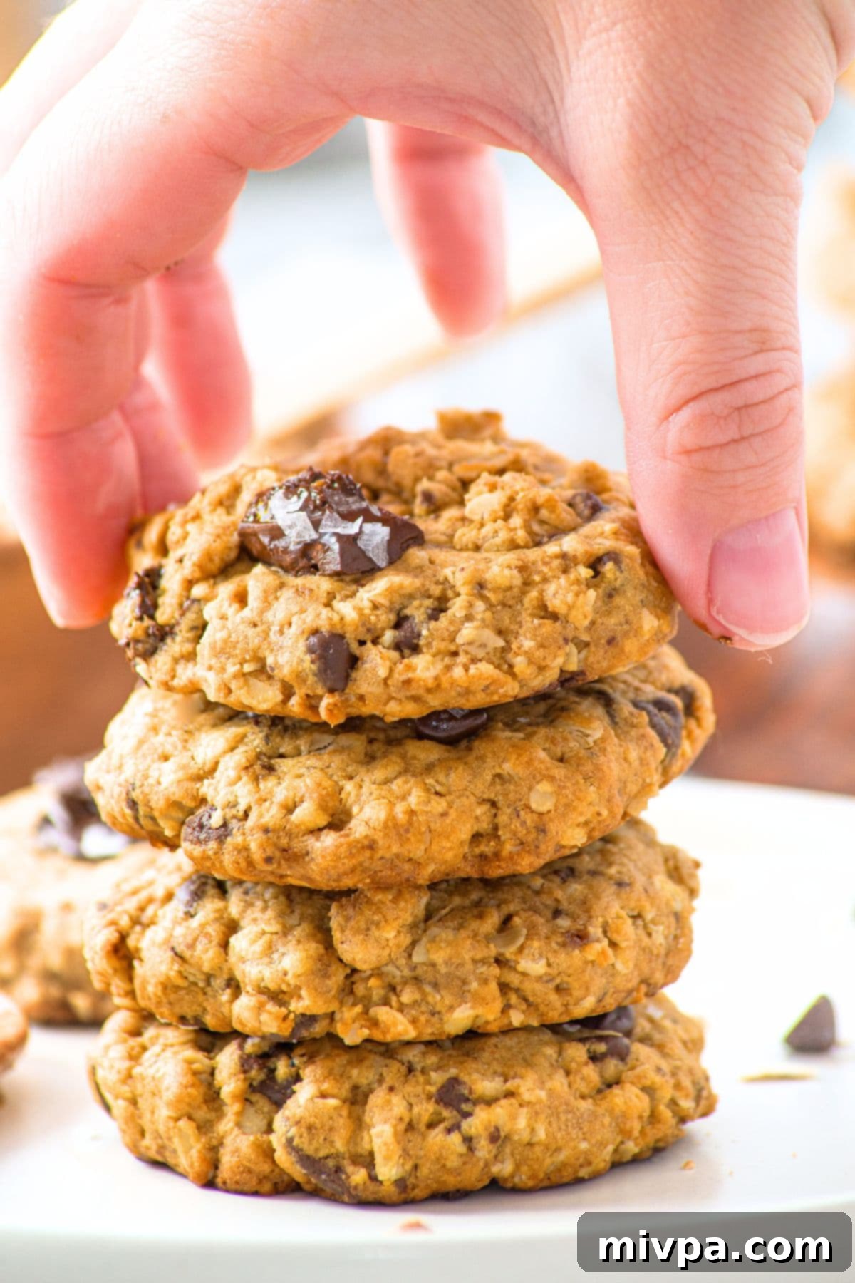 A hand reaching to pick up a single chocolate chip oatmeal cookie from a tall stack of freshly baked cookies.