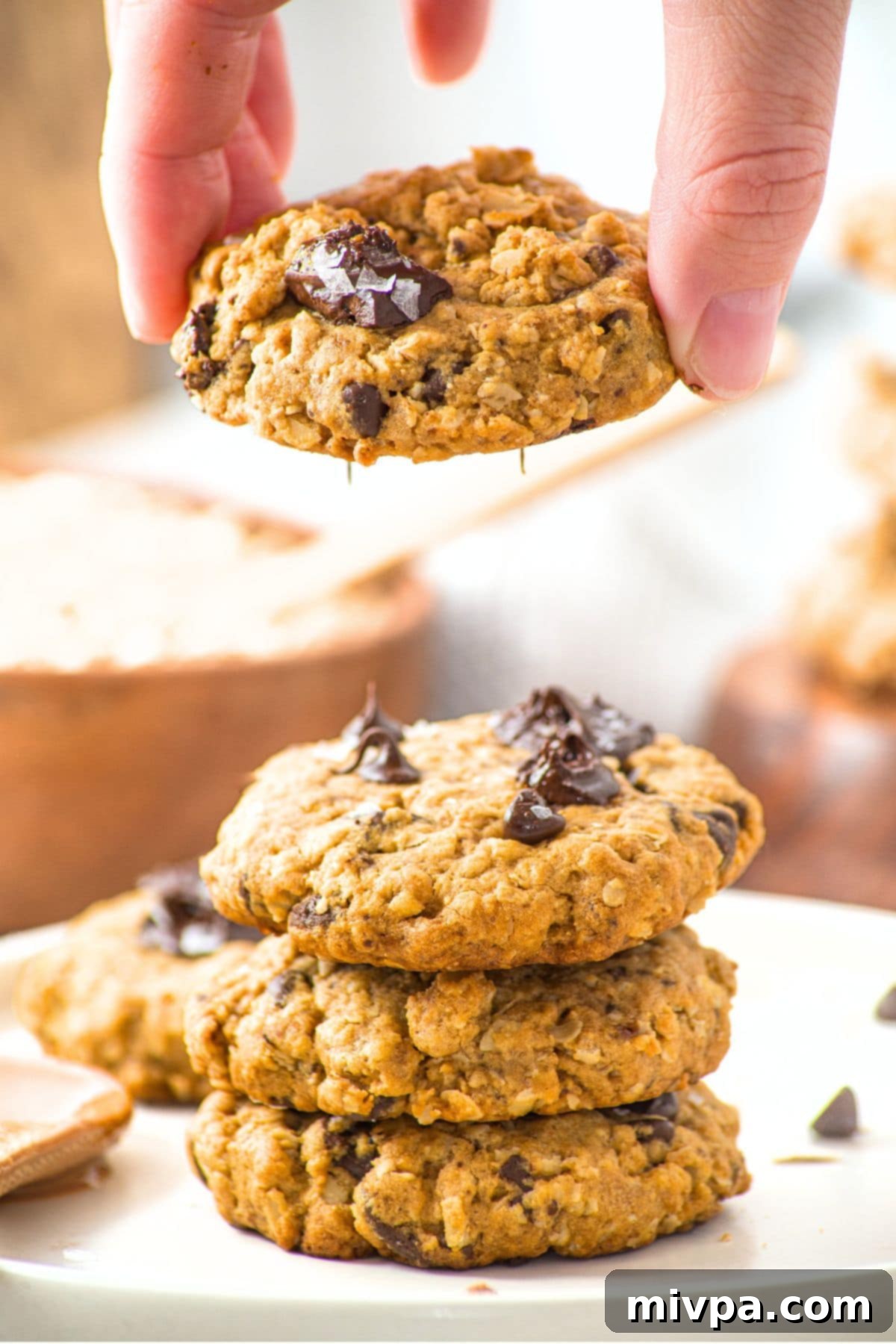 A hand holding up a freshly baked peanut butter oatmeal chocolate chip cookie, with a stack of cookies blurred in the background.
