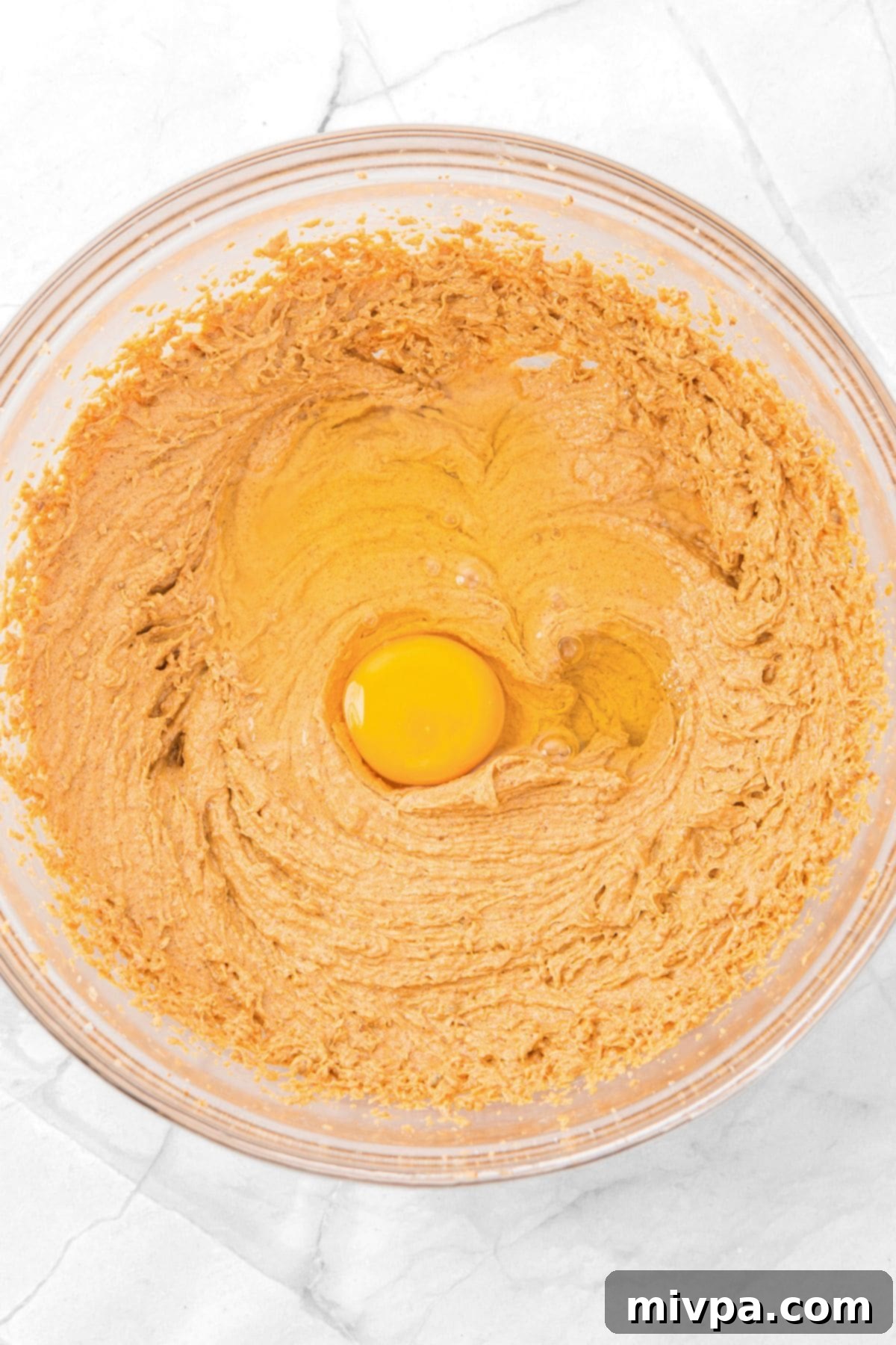 A single egg being cracked and dropped into the orange-brown batter in a glass mixing bowl.